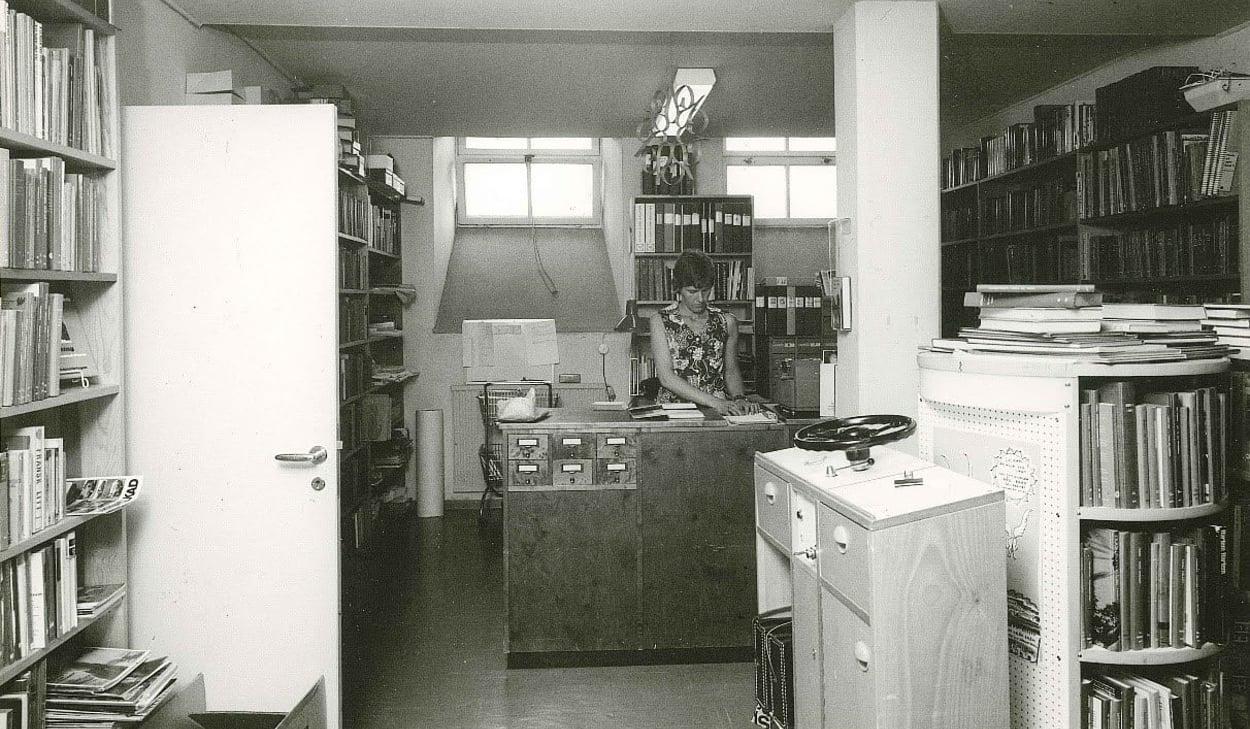 Svartvit bild av ett äldre bibliotek där en kvinna arbetar bakom en trädisk med kortkatalog. Rummet är fullt av bokhyllor, pärmar och dokument i ett trångt men organiserat utrymme.