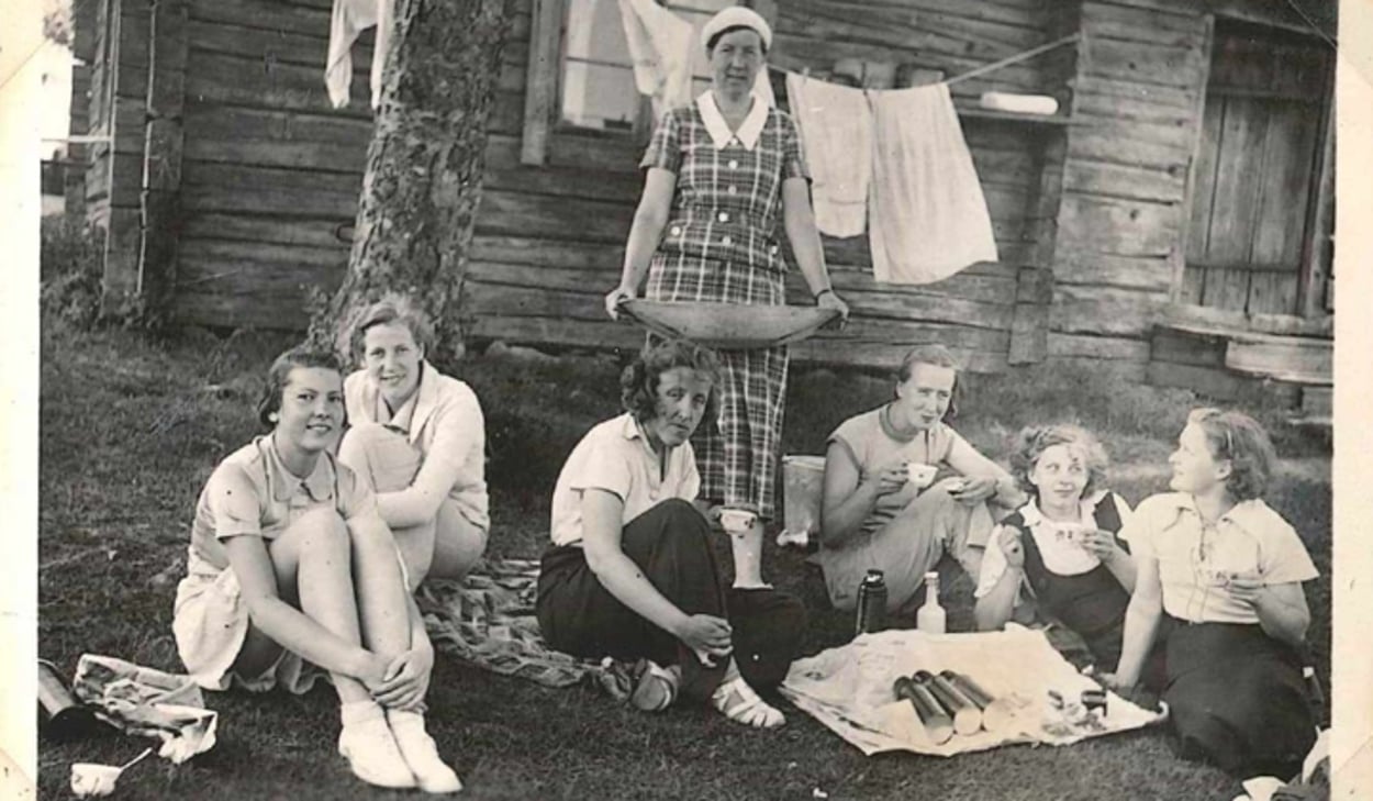Ett svartvitt foto av en grupp människor som sitter och fikar på gräset.