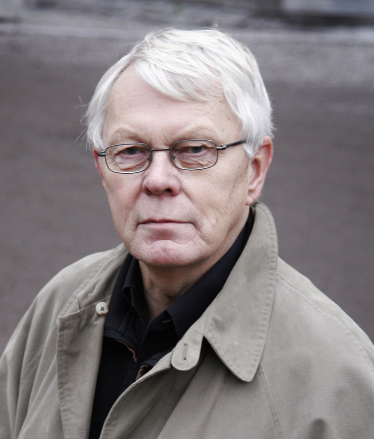 En man med vitt h&aring;r som b&auml;r en trenchcoat.