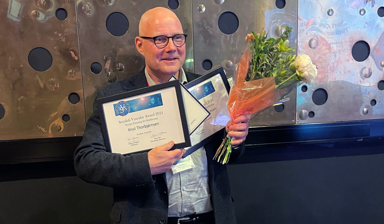 Kärlkirurgen Knut Thorbjørnsen, medelålders man står med blommor i handen och flera inramade prisdokument i handen. Han ler mot kameran. Han bär en ljus skjorta och en grå kavaj. 