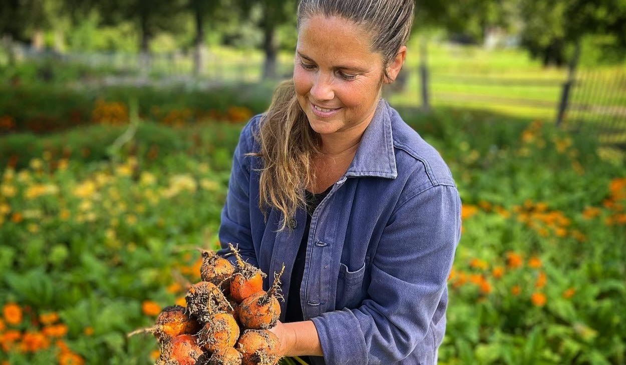 Person stående vid ett trädgårdsland och håller i grönsaker