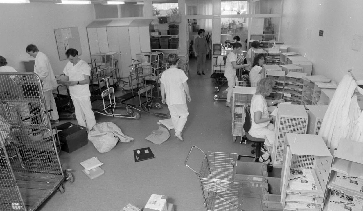 Gävle sjukhus transportavdelning den 5 maj 1989, fotograf är sjukhusfotografen Sven Östberg. I bild ser vi det organiserade kaoset när dagens post och paket ska transporteras ut till avdelningarna. Postsortering pågår vid postborden till höger, till vänster står sparkcyklarna redo att fyllas och i postfacken närmast i bild kan vi se, om vi tittar noga, att konstföreningens reklamblad är på väg att hitta ut till sina medlemmar.