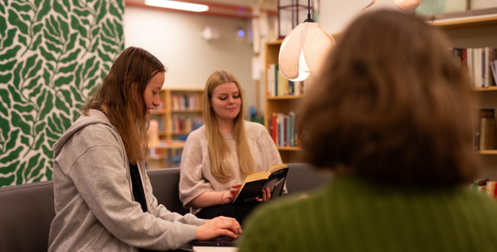Tre personer sitter i en soffa i ett bibliotek; en skriver på laptop, en läser en bok och den tredje är vänd bort från kameran.