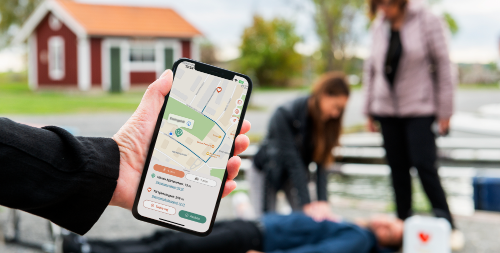 Hand med mobiltelefon, och i telefonen bild på en karta. I bakgrunden en suddig bild på tre personer, där hjärt-lungräddning pågår. Bilden är arrangerad och en kvinna står vid sidan, en annan ligger på marken. En tredje gör HLR. 