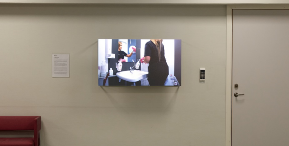 Skärm visar en videoinstallation med två personer vid ett handfat, uppsatt på en vägg i en korridor med bänk och stängd dörr.
