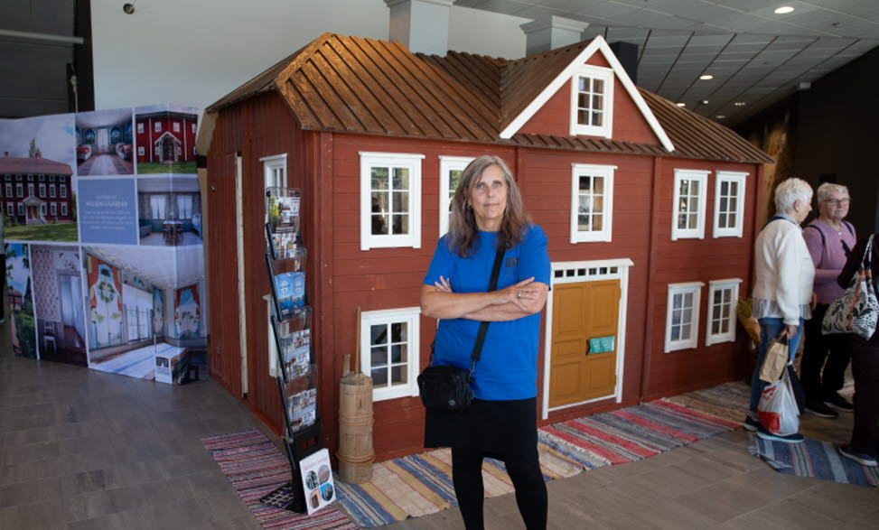 Kvinna i blå tröja står framför en röd modell av en traditionell Hälsingegård.