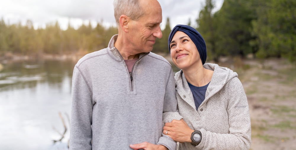 En pappa står och håller i sin vuxna dotter i armkrok. De tittar på varandra varmt och kärleksfullt. De står ute i naturen i en skog och en sjö skymtar i bakgrunden. 