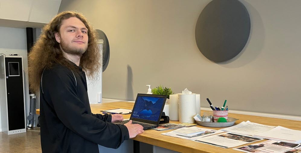 Person vid en dator ståendes vid en arbetsbänk med papper liggandes på sidan.